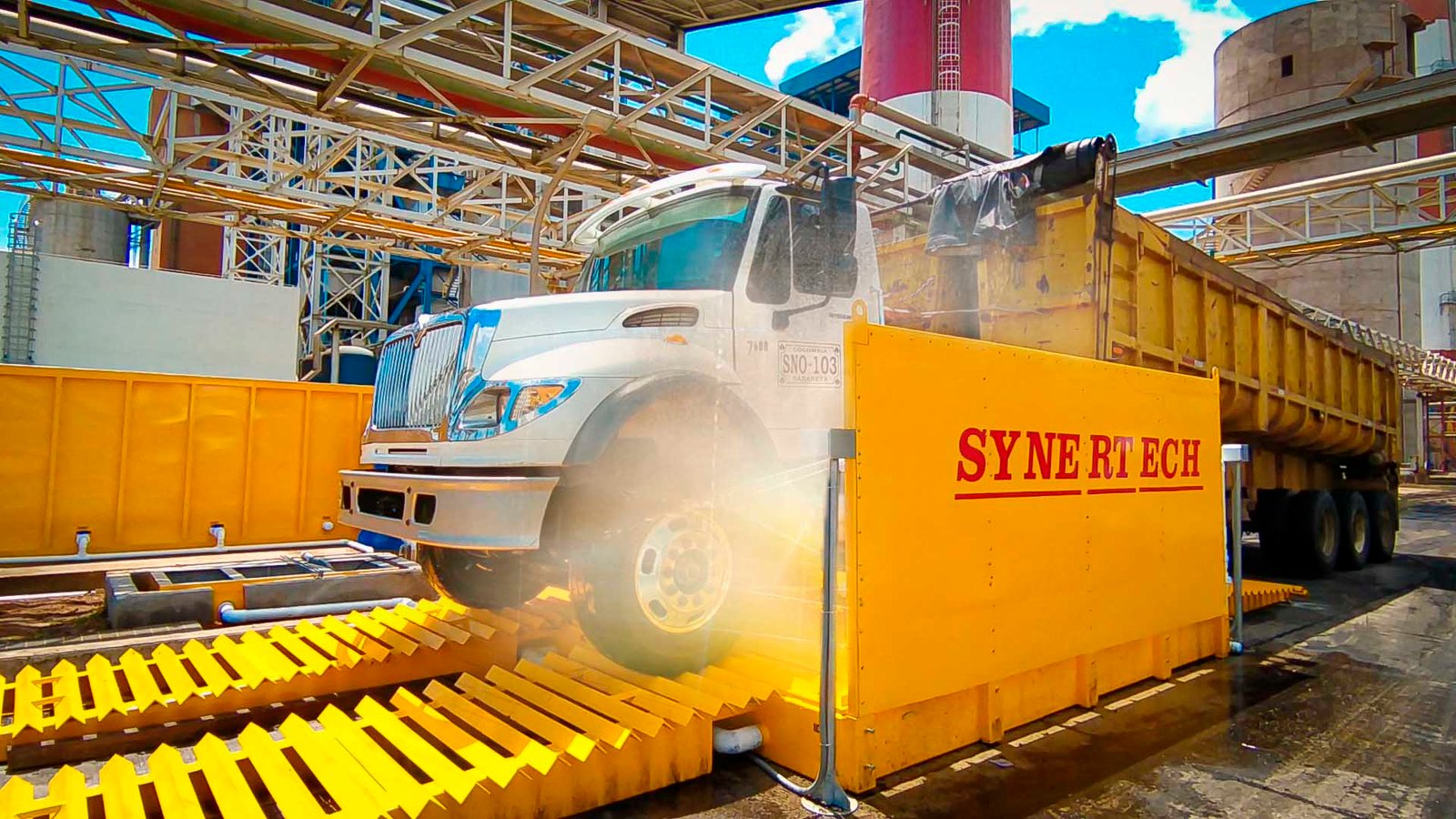 Camión grande blanco y amarillo pasando por un sistema de lavado de ruedas activo, rociando agua para limpiar la suciedad y escombros. En un sitio industrial o de construcción con silos al fondo, el sistema ayuda a mantener limpias las vías públicas. - ver más