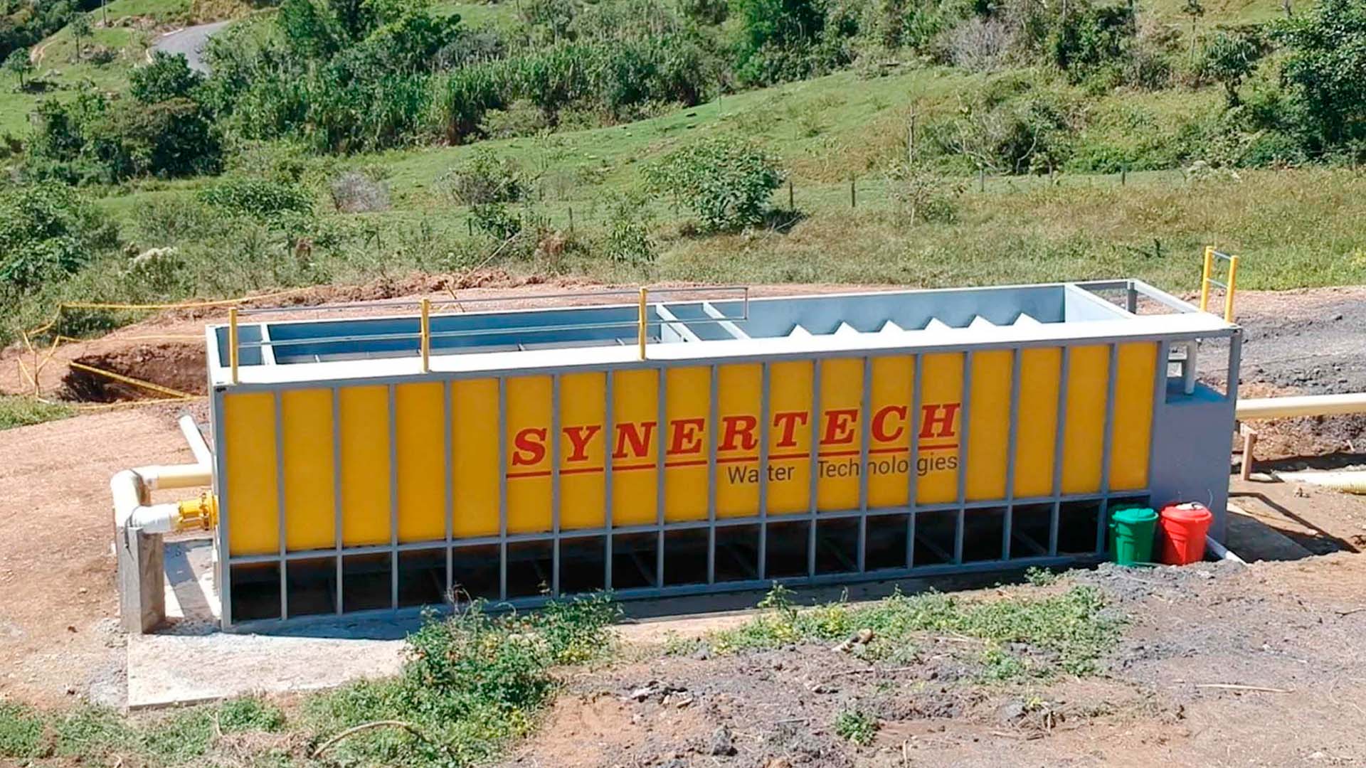 Camión rojo de plataforma larga transportando un gran decantador amarillo de la marca 'SYVERTECH' para el tratamiento de aguas residuales complejas, estacionado en una calle urbana con edificios y cielo despejado al fondo. - ver detalles