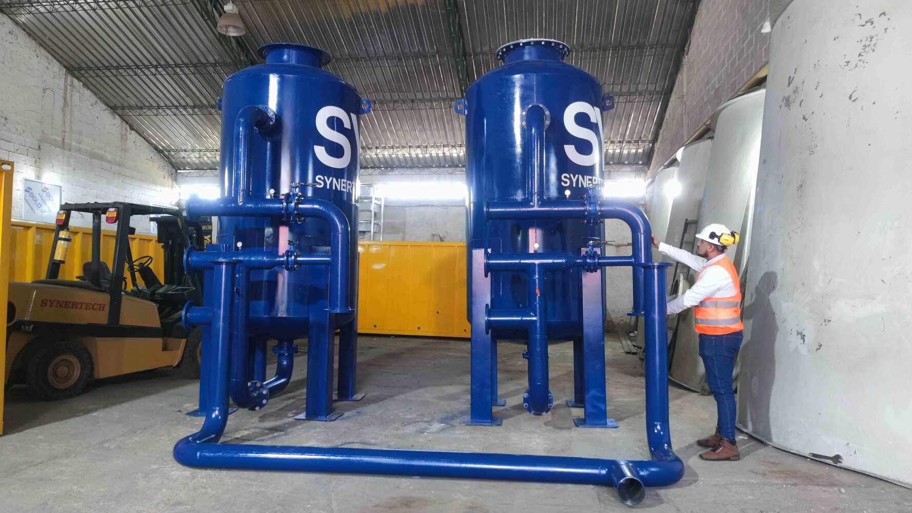 Técnico inspeccionando una planta de tratamiento de aguas residuales modular de color verde marca BIOMAX SYNERTECH, que incluye módulos de pretratamiento, decantación, DAF y oxidación avanzada. La planta está ubicada en un entorno industrial con techo alto y tuberías visibles. - ver más información